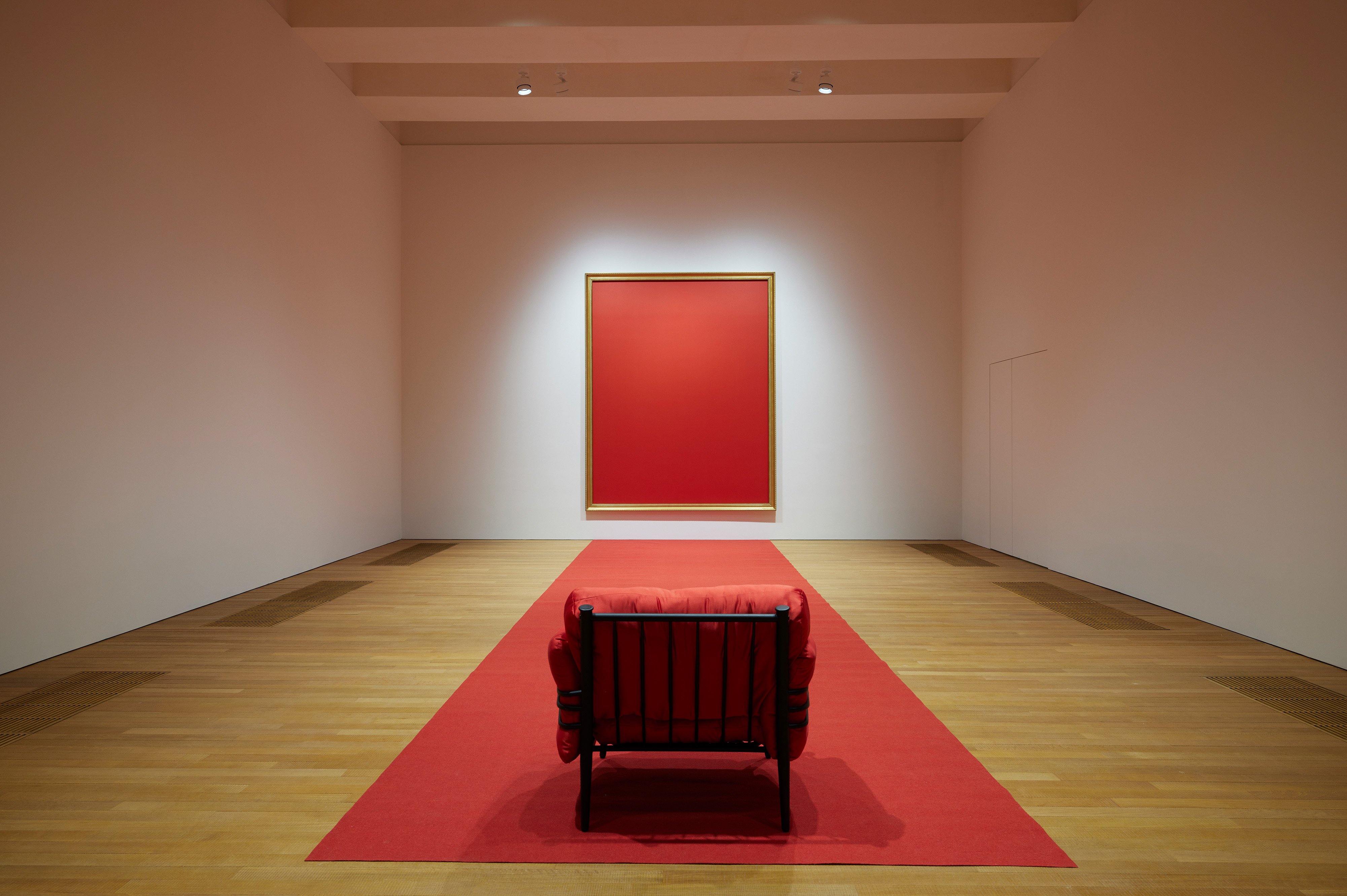 In a white-walled gallery with wooden flooring, a red-cushioned chair is placed facing the rear wall at the edge of a long red carpet. The carpet extends towards the rear wall, where a red painting in gilded frame is hung.